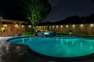 A backyard swimming pool and jacuzzi hot tob at night with solar lights around the fence for privacy and illumination.