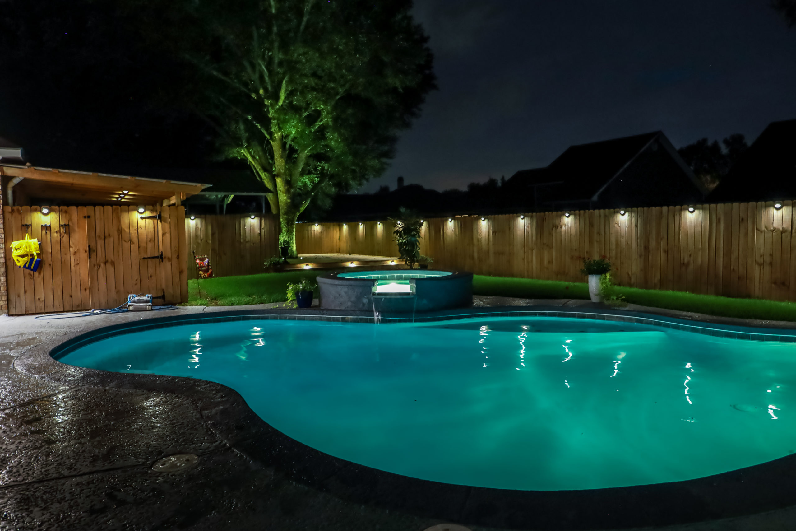 A backyard swimming pool and jacuzzi hot tob at night with solar lights around the fence for privacy and illumination.