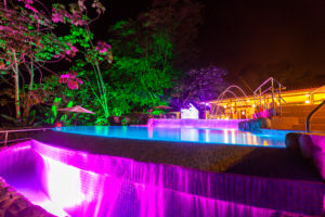 A calm swimming pool with glowing lights reflecting on the tropical trees around it with a darkened night sky in Costa Rica.