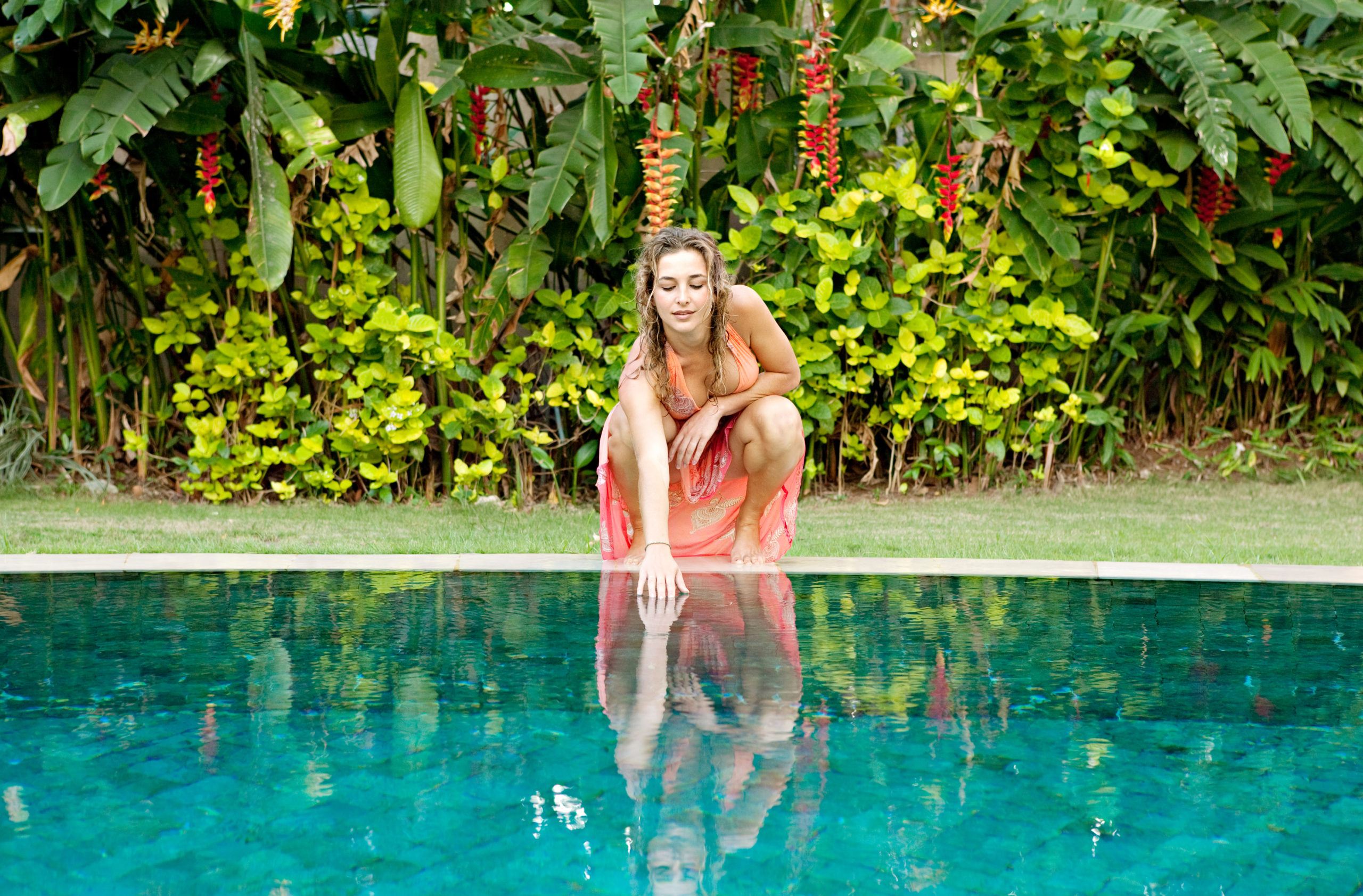 A woman reaches her hand into the pool water, surrounded by a backdrop of beautiful lush plants and foliage, enjoying a serene and nature-inspired moment by the pool.