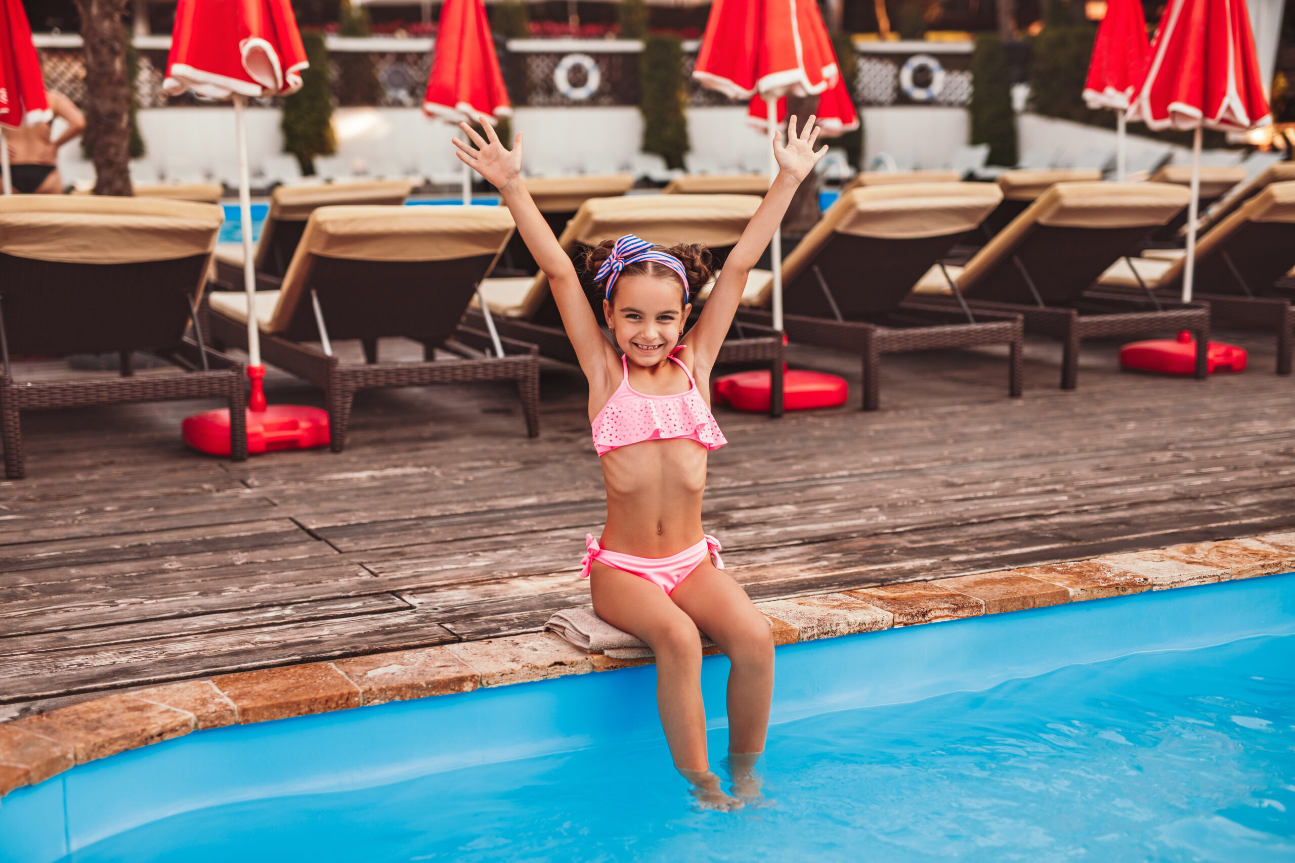 A child sitting at the edge of the pool with her arms raised in excitement, ready to jump or play in the water.